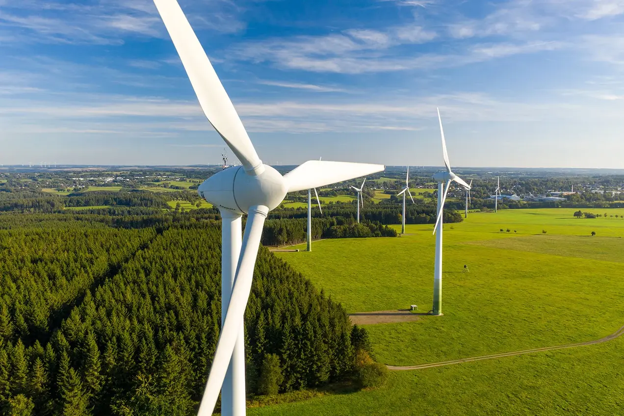 Luftaufnahme eines Windparks mit mehreren Windturbinen inmitten von grünen Wiesen und Wäldern. Der Himmel ist blau mit einigen Wolken, und die Landschaft erstreckt sich weitläufig in den Hintergrund.
