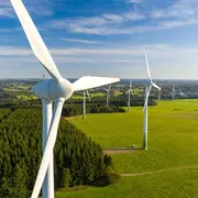 Luftaufnahme eines Windparks mit mehreren Windturbinen inmitten von grünen Wiesen und Wäldern. Der Himmel ist blau mit einigen Wolken, und die Landschaft erstreckt sich weitläufig in den Hintergrund.