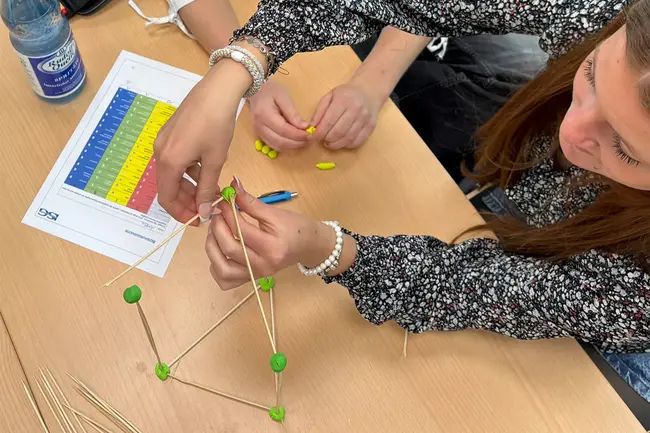 Eine Person baut mit Holzspießchen und bunten Knetkugeln eine Struktur auf einem Tisch. Auf dem Tisch liegt ein farbiges Blatt Papier. Die Person konzentriert sich auf die Konstruktion, während eine zweite Hand ebenfalls Teil des kreativen Prozesses ist.