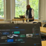 Ein Referent steht an einem Tisch und präsentiert vor einer Gruppe. Auf dem Tisch liegt ein Laptop mit einer Präsentationsfolie über Batteriesysteme. Im Hintergrund sind Fenster mit Blick auf Bäume sichtbar.