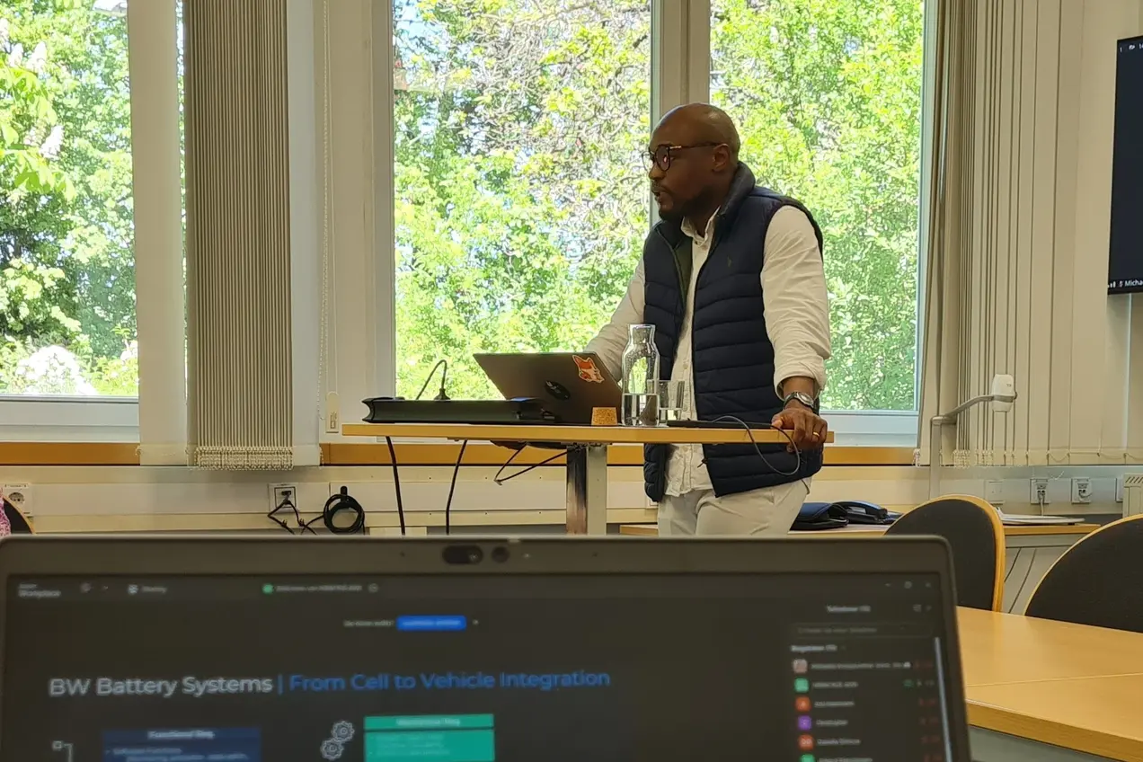 Ein Referent steht an einem Tisch und präsentiert vor einer Gruppe. Auf dem Tisch liegt ein Laptop mit einer Präsentationsfolie über Batteriesysteme. Im Hintergrund sind Fenster mit Blick auf Bäume sichtbar.