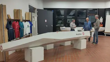 In einem modernen Ausstellungsraum stehen drei Personen vor einem länglichen Boot, das auf einem Ständer platziert ist. Im Hintergrund sind Sportshirts und eine Leinwand sichtbar. Die Atmosphäre ist informativ und ansprechend.