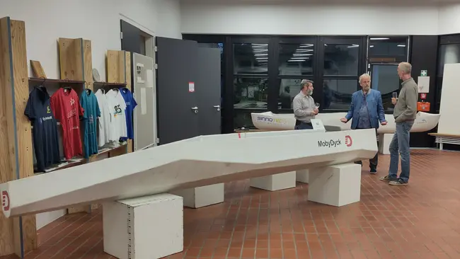 In einem modernen Ausstellungsraum stehen drei Personen vor einem länglichen Boot, das auf einem Ständer platziert ist. Im Hintergrund sind Sportshirts und eine Leinwand sichtbar. Die Atmosphäre ist informativ und ansprechend.