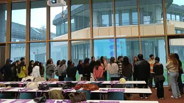 Eine Gruppe von etwa 20 Personen steht in einem modernen Raum vor großen Fenstern. Sie diskutieren, während im Hintergrund ein Gebäude zu sehen ist. Auf den Tischen liegen persönliche Gegenstände und Informationsmaterialien. Es herrscht eine freundliche, kommunikative Atmosphäre.