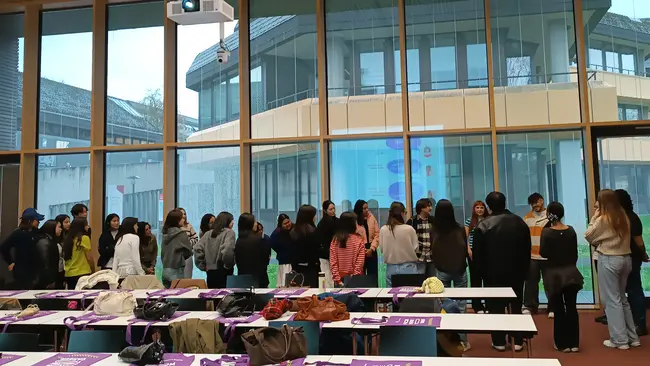 Eine Gruppe von etwa 20 Personen steht in einem modernen Raum vor großen Fenstern. Sie diskutieren, während im Hintergrund ein Gebäude zu sehen ist. Auf den Tischen liegen persönliche Gegenstände und Informationsmaterialien. Es herrscht eine freundliche, kommunikative Atmosphäre.