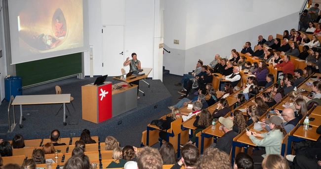 Eine Person steht an einem Podium in einem Hörsaal und präsentiert vor einem Publikum. Im Hintergrund ist eine Projektionsleinwand zu sehen, auf der ein Bild angezeigt wird. Das Publikum besteht aus zahlreichen Studierenden, die aufmerksam zuhören.