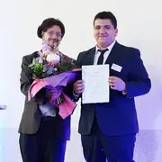 Anes Khaldi hält ein Dokument in der Hand und lächelt. Neben ihm steht Prof. Dr. Christian Schachtner, der einen Blumenstrauß hält. 