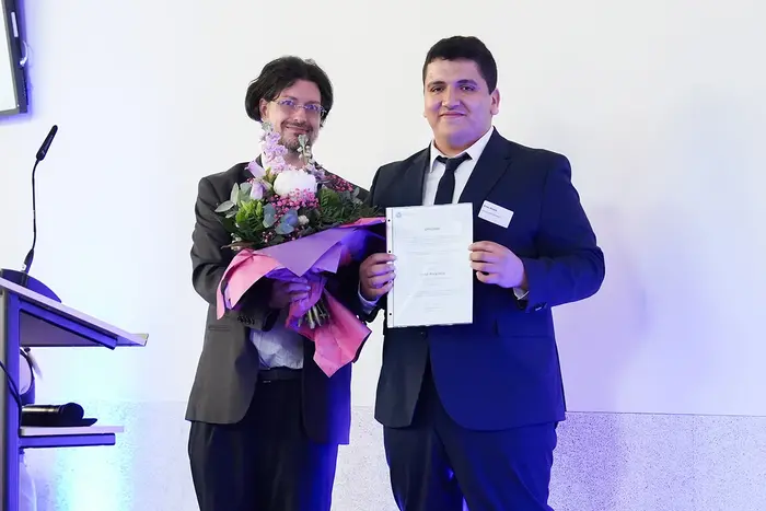 Anes Khaldi hält ein Dokument in der Hand und lächelt. Neben ihm steht Prof. Dr. Christian Schachtner, der einen Blumenstrauß hält. 