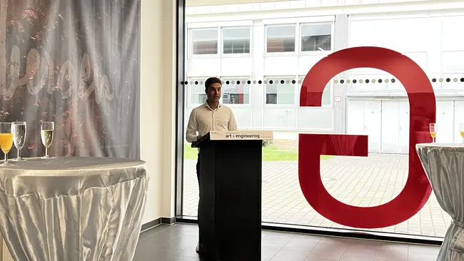 Ein Mann steht an einem Rednerpult in einem Raum mit großen Fenstern. Im Hintergrund ist ein Banner zu sehen, das den Schriftzug "erlangen" zeigt. Auf den Tischen stehen Sektgläser. Die Atmosphäre wirkt festlich und einladend.