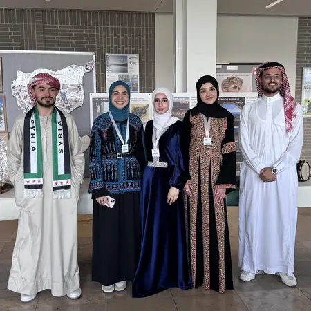 Fünf Personen stehen nebeneinander in traditioneller arabischer Kleidung. Zwei Frauen tragen lange, farbenfrohe Gewänder, während die Männer in weißen und hellen Gewändern gekleidet sind. Hintergrund zeigt kulturelle Ausstellungsstücke. Alle Personen lächeln.