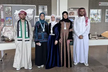 Fünf Personen stehen nebeneinander in traditioneller arabischer Kleidung. Zwei Frauen tragen lange, farbenfrohe Gewänder, während die Männer in weißen und hellen Gewändern gekleidet sind. Hintergrund zeigt kulturelle Ausstellungsstücke. Alle Personen lächeln.
