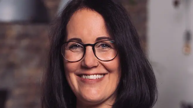 Eine lächelnde Frau mit schulterlangen, dunklen Haaren trägt eine graue Blazerjacke über einem weißen T-Shirt. Sie steht vor einem hellen Hintergrund und wirkt freundlich und professionell. Ihre Brille verstärkt den positiven Eindruck.