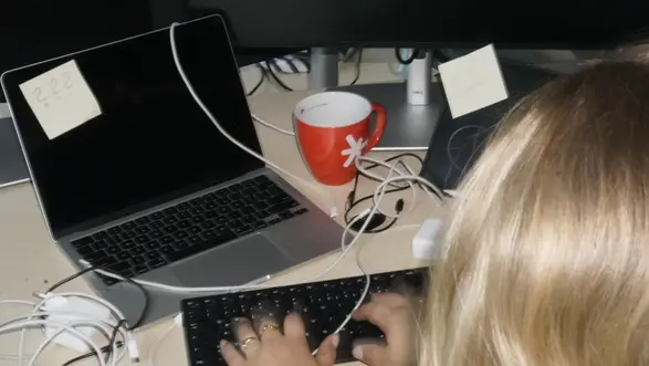 Eine Person mit langen, blonden Haaren tippt auf einer schmalen Tastatur, während sie am Computer arbeitet. Neben dem Laptop steht eine rote Tasse. An den Bildschirmen sind Notizzettel befestigt. Kabel sind verstreut auf dem Tisch.