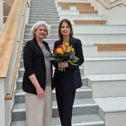 Auf der Treppe stehen zwei Frauen. Prof. Dr. Ursula Walkenhorst trägt eine schwarze Jacke, während Prof. Dr. Susanne Rägle einen dunklen Anzug trägt und einen bunten Blumenstrauß hält. Beide lächeln und stehen freundlich zusammen.