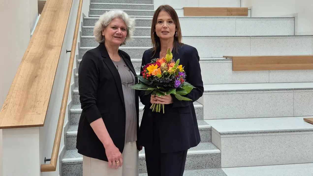Auf der Treppe stehen zwei Frauen. Prof. Dr. Ursula Walkenhorst trägt eine schwarze Jacke, während Prof. Dr. Susanne Rägle einen dunklen Anzug trägt und einen bunten Blumenstrauß hält. Beide lächeln und stehen freundlich zusammen.