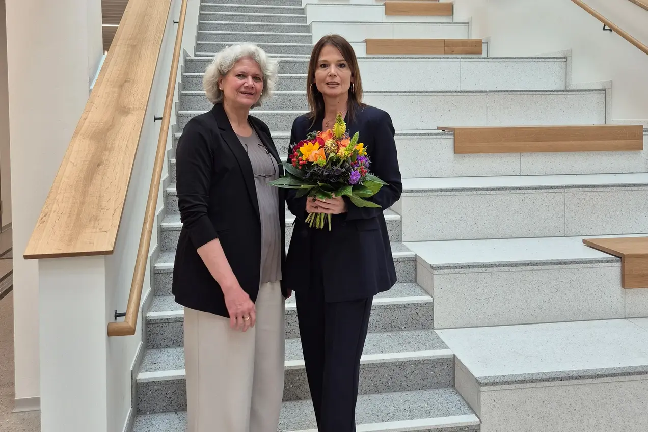 Two women are standing on the stairs. Prof. Dr. Ursula Walkenhorst is wearing a black jacket, while Prof. Dr. Susanne Rägle is wearing a dark suit and holding a colorful bouquet of flowers. Both are smiling and standing together in a friendly manner.