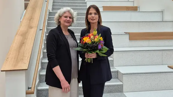 Auf der Treppe stehen zwei Frauen. Prof. Dr. Ursula Walkenhorst trägt eine schwarze Jacke, während Prof. Dr. Susanne Rägle einen dunklen Anzug trägt und einen bunten Blumenstrauß hält. Beide lächeln und stehen freundlich zusammen.