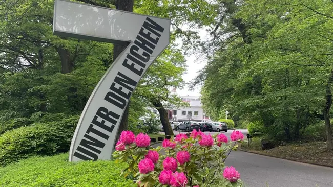 Ein großes, schräges Schild mit der Aufschrift "UNTER DEN EICHEN" steht am Eingang eines Weges. Um das Schild blühen pinkfarbene Rhododendren in einem grünen Bereich. Im Hintergrund sind Bäume sichtbar, die eine natürliche Umgebung schaffen.