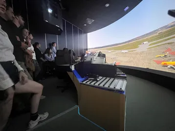Das Bild zeigt eine Gruppe von Menschen, die in einem Simulator stehen und auf eine große, gebogene Leinwand schauen, die einen Flughafen mit landenden und startenden Flugzeugen zeigt. Vor ihnen befindet sich ein Kontrollpult mit Bedienelementen. Die Atmosphäre ist konzentriert und lehrreich.