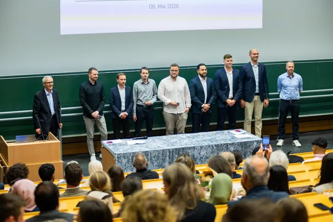 Eine Gruppe von acht männlichen Personen steht auf einer Bühne in einem Hörsaal. Vor ihnen befindet sich ein Tisch mit einem grauen Tuch. Im Publikum sitzen Zuschauer, die auf die Bühne schauen. Im Hintergrund sind Lehrstühle sichtbar. Das Datum auf der Leinwand ist der 6. Mai 2026.