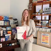 Eine lächelnde Frau in einem hellen Anzug hält ein Dokument mit der Aufschrift "We train the best!" in der Hand. Im Hintergrund sind Regale mit verschiedenen Büchern und Gegenständen zu sehen, die eine freundliche Atmosphäre schaffen.