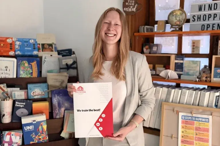 Eine lächelnde Frau in einem hellen Anzug hält ein Dokument mit der Aufschrift "We train the best!" in der Hand. Im Hintergrund sind Regale mit verschiedenen Büchern und Gegenständen zu sehen, die eine freundliche Atmosphäre schaffen.