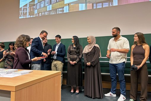 Absolvent:innen der Hochschule RheinMain stehen in einer Reihe und erhalten nacheinander ein Präsent aus den Händen der Vorsitzenden der Prüfungsausschüsse