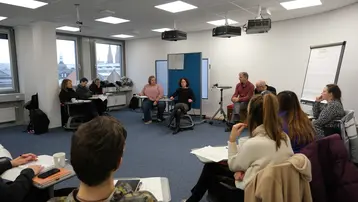 Eine Gruppenbesprechung in einem modernen Seminarraum. Teilnehmer sitzen in einem Halbkreis, während mehrere Sprecher auf Stühlen in der Mitte sitzen. Sie diskutieren, während Notizblöcke und Getränke auf den Tischen vor den Zuhörern liegen. Große Fenster im Hintergrund bieten Tageslicht.