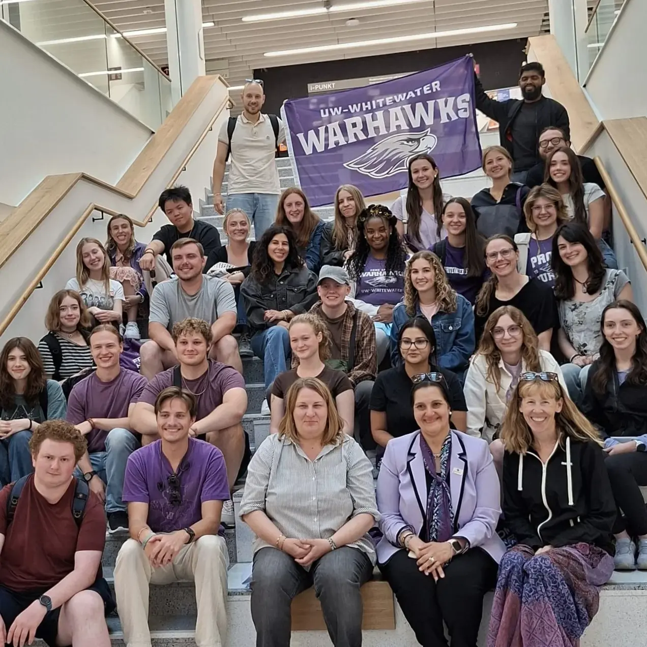 Eine Gruppe von etwa 40 Personen sitzt und steht auf einer Treppe in einem modernen Gebäude. Viele tragen Kleidung in lila und schwarz, und im Hintergrund hängt ein Banner mit der Aufschrift "UW-Whitewater Warthawks". Alle schauen in die Kamera und lächeln.