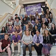Eine Gruppe von etwa 40 Personen sitzt und steht auf einer Treppe in einem modernen Gebäude. Viele tragen Kleidung in lila und schwarz, und im Hintergrund hängt ein Banner mit der Aufschrift "UW-Whitewater Warthawks". Alle schauen in die Kamera und lächeln.