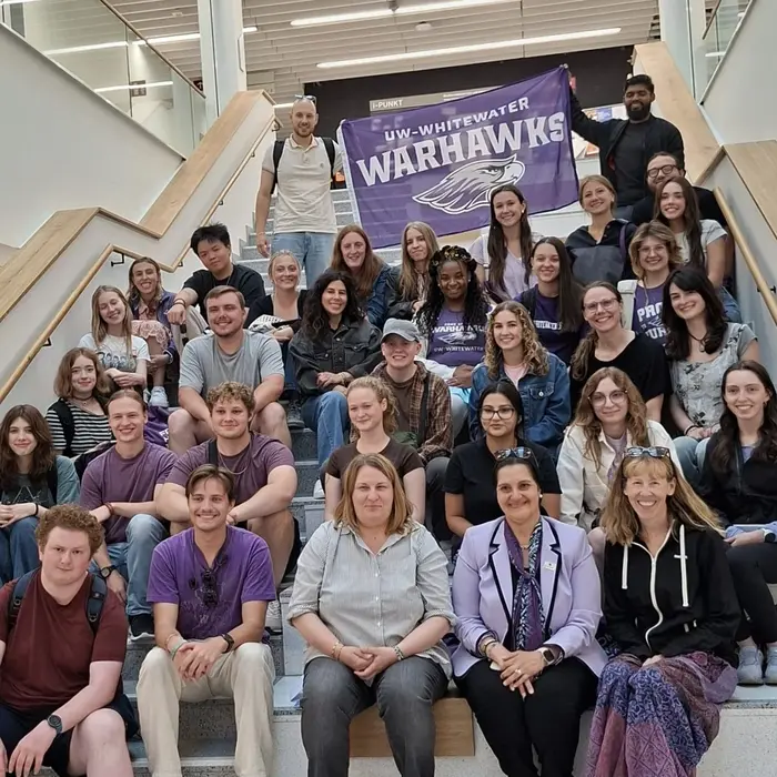 Eine Gruppe von etwa 40 Personen sitzt und steht auf einer Treppe in einem modernen Gebäude. Viele tragen Kleidung in lila und schwarz, und im Hintergrund hängt ein Banner mit der Aufschrift "UW-Whitewater Warthawks". Alle schauen in die Kamera und lächeln.