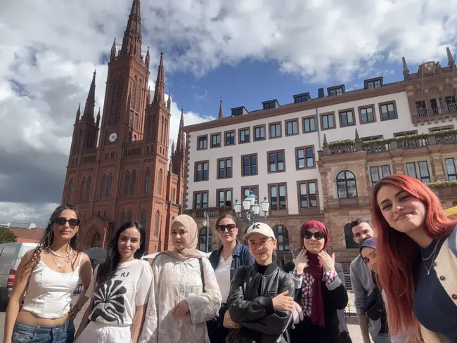 Eine Gruppe von acht Personen steht lächelnd vor einem großen, roten historischen Gebäude mit einer Turmuhr. Im Hintergrund sind moderne Gebäude und ein bewölkter Himmel zu sehen. Die Menschen zeigen eine freundliche, einladende Haltung.