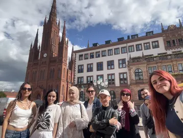 Eine Gruppe von acht Personen steht lächelnd vor einem großen, roten historischen Gebäude mit einer Turmuhr. Im Hintergrund sind moderne Gebäude und ein bewölkter Himmel zu sehen. Die Menschen zeigen eine freundliche, einladende Haltung.