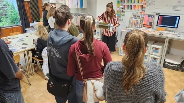 Eine Gruppe von Personen steht in einem Raum und sieht einer Präsentation zu. Eine Frau mit gestreiftem Oberteil zeigt Materialien und spricht. Auf einem Tisch stehen verschiedene Papiere und ein Computer mit Bildschirm. Die Stimmung ist aufmerksam und interessiert.