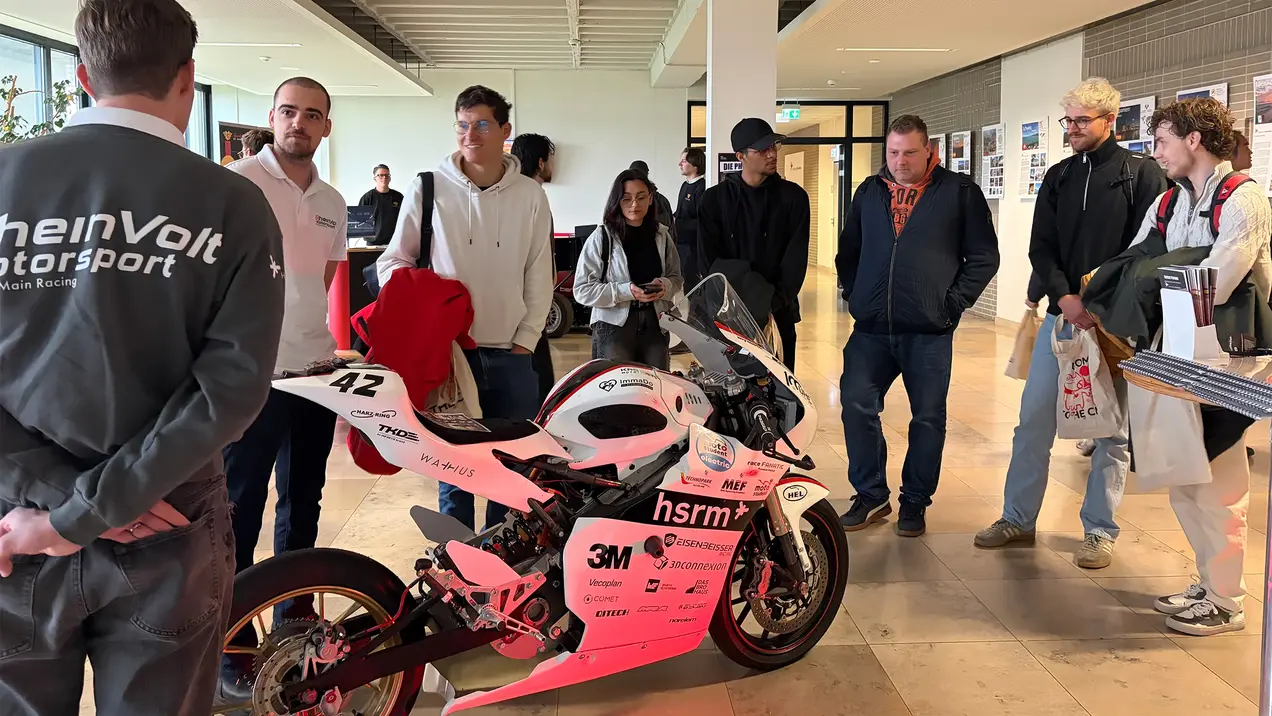 Eine Gruppe von Menschen betrachtet ein sportliches Motorrad mit der Nummer 42. Es steht im Vordergrund, während eine Person im grauen Outfit mit dem Logo „RheinVolt Motorsport“ erklärt. Im Hintergrund sind weitere Besucher und informative Displays zu sehen.