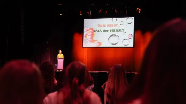 Eine Person steht auf einer Bühne und präsentiert eine Folie mit der Frage „Was ist dann der AStA der HSRM?“ Im Hintergrund sind Zuschauer zu sehen, die auf den Vortrag achten. Die Bühne ist beleuchtet, und es gibt ein visuelles Element mit Blasen.