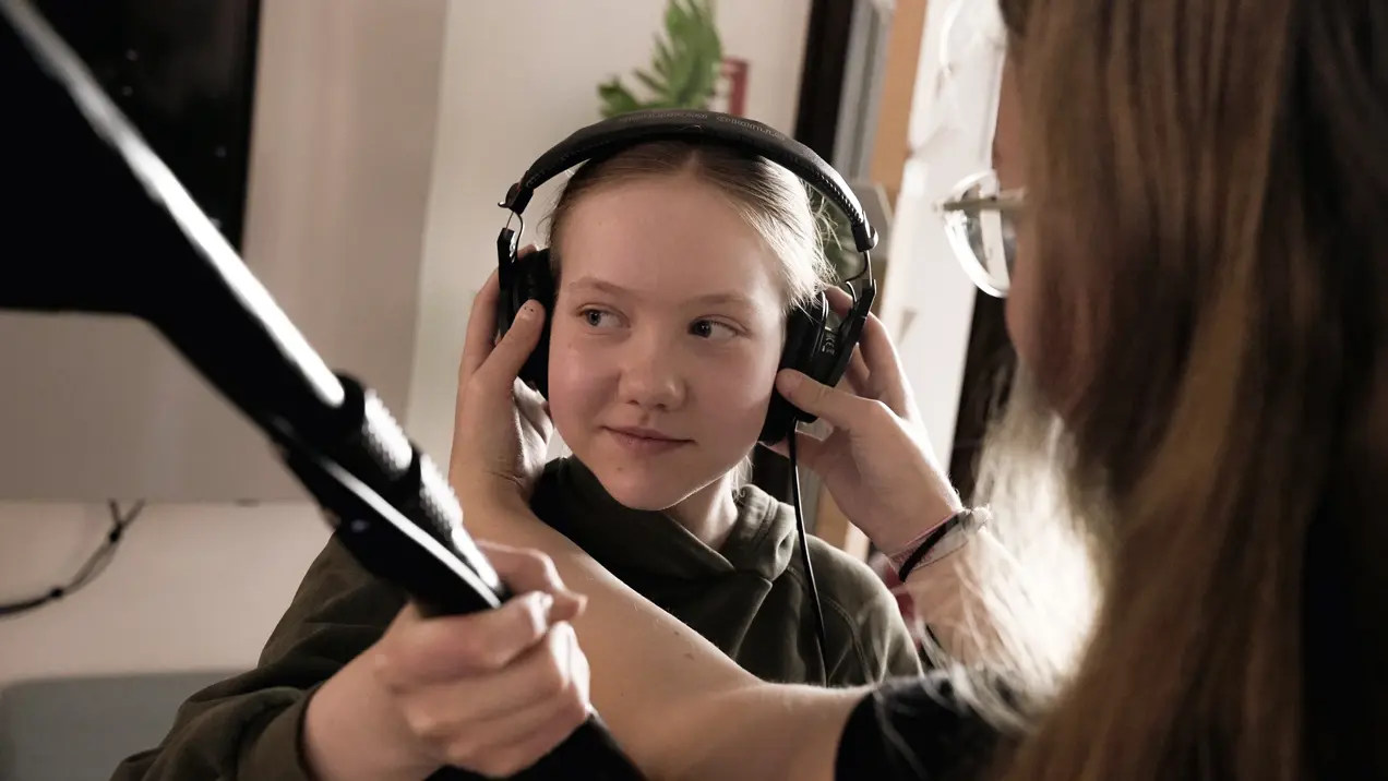 Ein Mädchen trägt Kopfhörer und wird von einer anderen Person, die ihr bei der Handhabung hilft, betreut. Die Szene vermittelt einen Moment der Aufmerksamkeit und des Austauschs während einer Aufnahme oder Aufnahmeprüfung.
