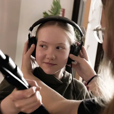 Ein Mädchen trägt Kopfhörer und wird von einer anderen Person, die ihr bei der Handhabung hilft, betreut. Die Szene vermittelt einen Moment der Aufmerksamkeit und des Austauschs während einer Aufnahme oder Aufnahmeprüfung.