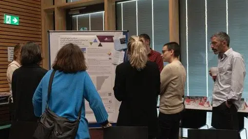 Eine Gruppe von sieben Personen steht um ein großes Poster, das Informationen und Grafiken enthält. Die Anwesenden diskutieren lebhaft, während sie auf verschiedene Aspekte des Posters zeigen. Der Raum hat Holzverkleidungen und große Fenster, die natürliches Licht hereinlassen.