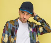 Der Autor Leif Randt mit bunter Jacke und Basecap vor einem gelben Hintergrund
