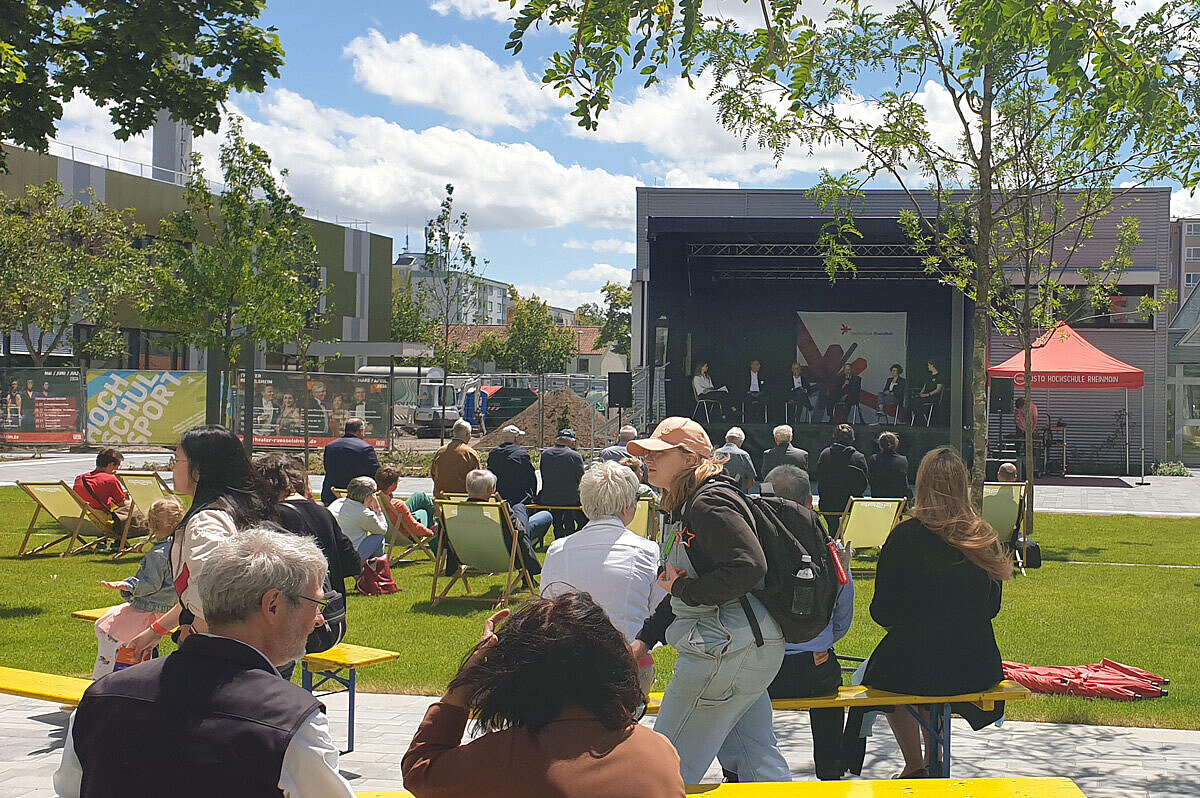 Besucher:innen des Campus Rüsselsheim sitzen auf Bänken und Stühlen vor einer Bühne 