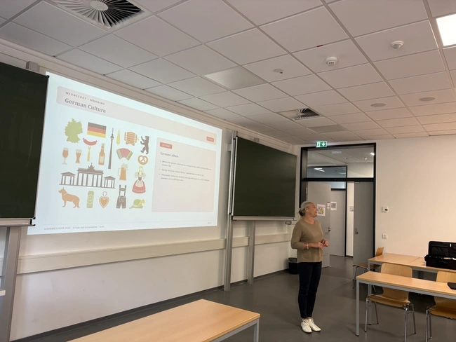 Eine Frau steht in einem Klassenzimmer und präsentiert Informationen über die deutsche Kultur. Auf der Leinwand sind verschiedene Symbole und Bilder dargestellt, die deutsche Traditionen und Sehenswürdigkeiten repräsentieren. Der Raum ist mit Tischen und Stühlen ausgestattet.