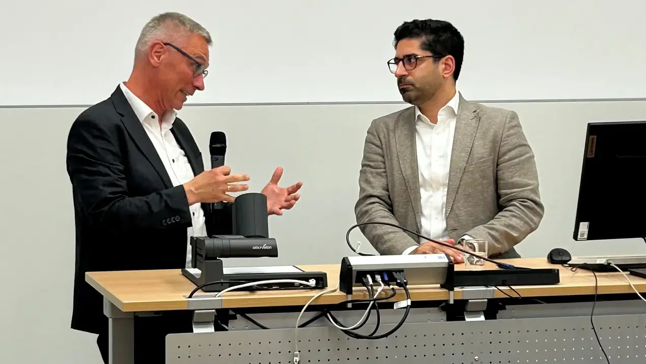 Zwei Männer stehen an einem Tisch in einem Präsentationsraum. Der Mann links hält ein Mikrofon und spricht, während der Mann rechts aufmerksam zuhört. Auf dem Tisch befindet sich technisches Equipment, darunter ein Computer und ein Projektor.