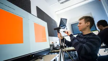 Ein junger Mann justiert ein Mikrofon vor einem Bildschirm, auf dem zwei orangefarbene Rechtecke angezeigt werden. Im Hintergrund arbeitet eine weitere Person am Computer. Der Raum wirkt modern und technisch eingerichtet.