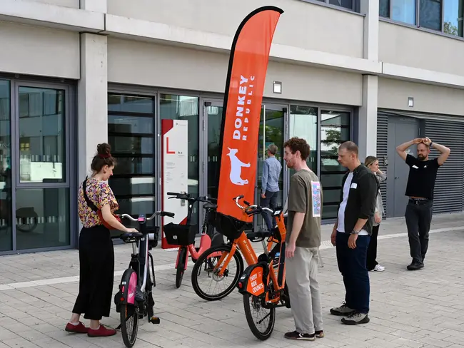 Die Bildbeschreibung zeigt eine Gruppe von Personen, die an einem Fahrradverleihstand stehen. Eine Frau und ein Mann betrachten zwei Fahrräder, während ein anderer Mann daran interessiert ist. Im Hintergrund weht ein rotes Banner mit dem Logo eines Fahrradverleihs.