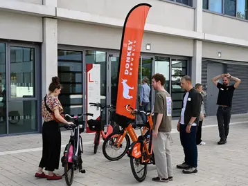 Die Bildbeschreibung zeigt eine Gruppe von Personen, die an einem Fahrradverleihstand stehen. Eine Frau und ein Mann betrachten zwei Fahrräder, während ein anderer Mann daran interessiert ist. Im Hintergrund weht ein rotes Banner mit dem Logo eines Fahrradverleihs.