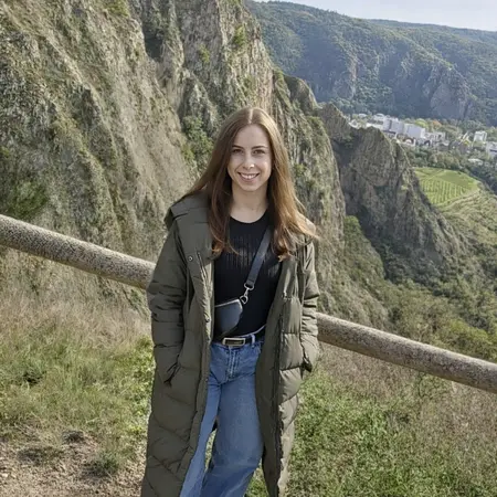 Eine junge Frau steht an einem Aussichtspunkt mit einem beeindruckenden Bergpanorama im Hintergrund. Sie trägt einen langen, grünen Mantel und Jeans, lächelt freundlich und steht neben einem Holzgeländer, während die Landschaft mit Hügeln und einer Stadt im Hintergrund sichtbar ist.