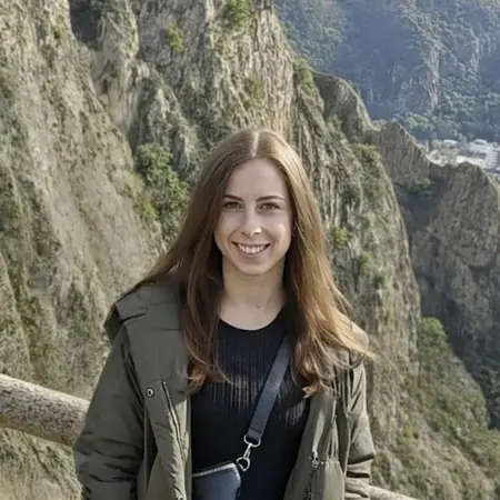 Eine junge Frau steht an einem Aussichtspunkt mit einem beeindruckenden Bergpanorama im Hintergrund. Sie trägt einen langen, grünen Mantel und Jeans, lächelt freundlich und steht neben einem Holzgeländer, während die Landschaft mit Hügeln und einer Stadt im Hintergrund sichtbar ist.