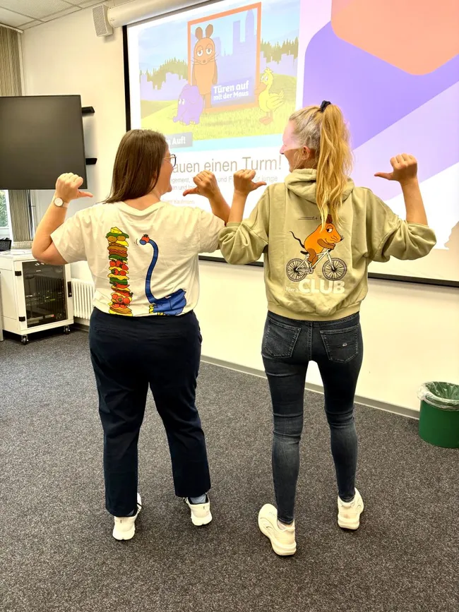Zwei Frauen stehen Rücken an Rücken. Eine trägt ein T-Shirt mit einem bunten Motiv eines Monsters, die andere einen Hoodie mit einem Cartoon-Hasen. Beide zeigen mit den Daumen auf sich selbst. Im Hintergrund ist ein Präsentationsbildschirm mit einer farbigen Grafik zu sehen.