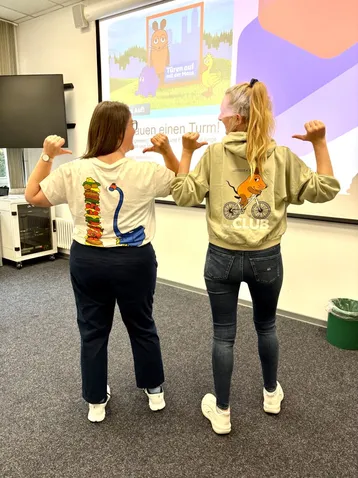 Zwei Frauen stehen Rücken an Rücken. Eine trägt ein T-Shirt mit einem bunten Motiv eines Monsters, die andere einen Hoodie mit einem Cartoon-Hasen. Beide zeigen mit den Daumen auf sich selbst. Im Hintergrund ist ein Präsentationsbildschirm mit einer farbigen Grafik zu sehen.